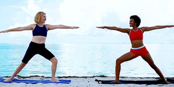 Outplay gender neutral swimwear and activewear - models in warrior pose on the sand at the beach. Model on the right is wearing a red compression swim top Swimmee and red Tomboier swim shorts. Model on the left is wearing a navy blue compression swim top Swimmee and navy blue and black Medina jammers.